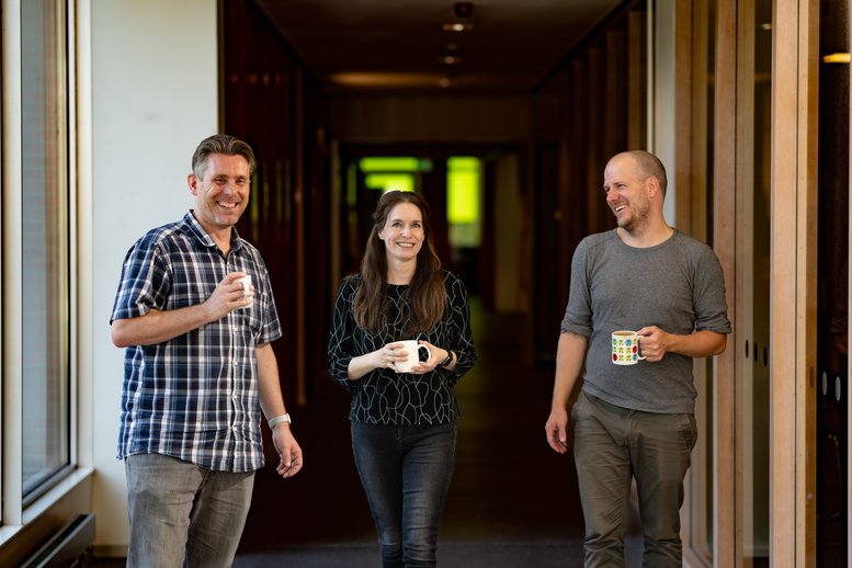 Three researchers in the hallway, one woman and two men, holding coffee mugs.